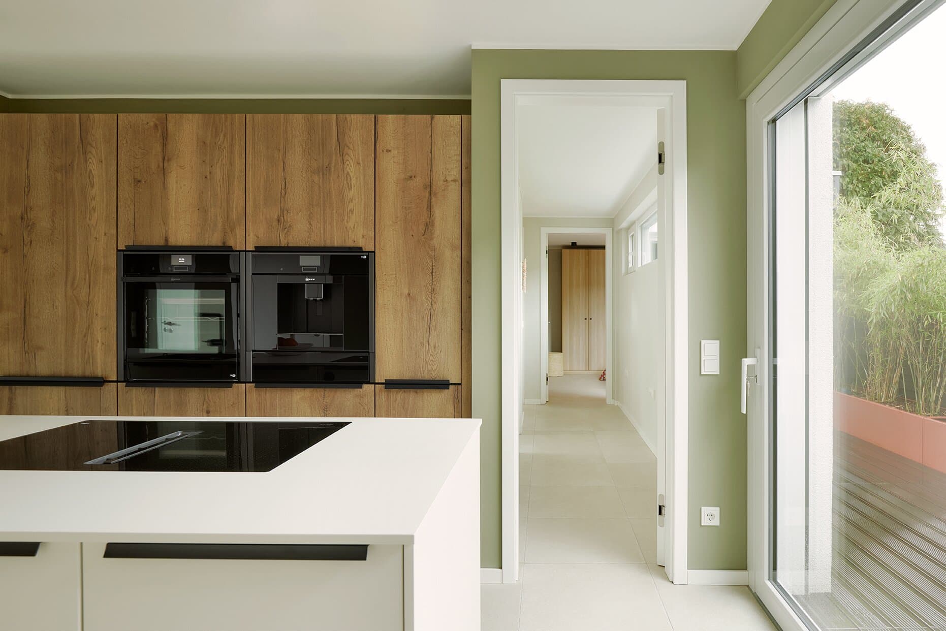 Kücheninsel mit Induktionskochfeld, Eichen-Hochschrankzeile mit Backofen und integriertem Kaffeevollautomat, salbeigrüne Wand und Blick auf die Terrasse