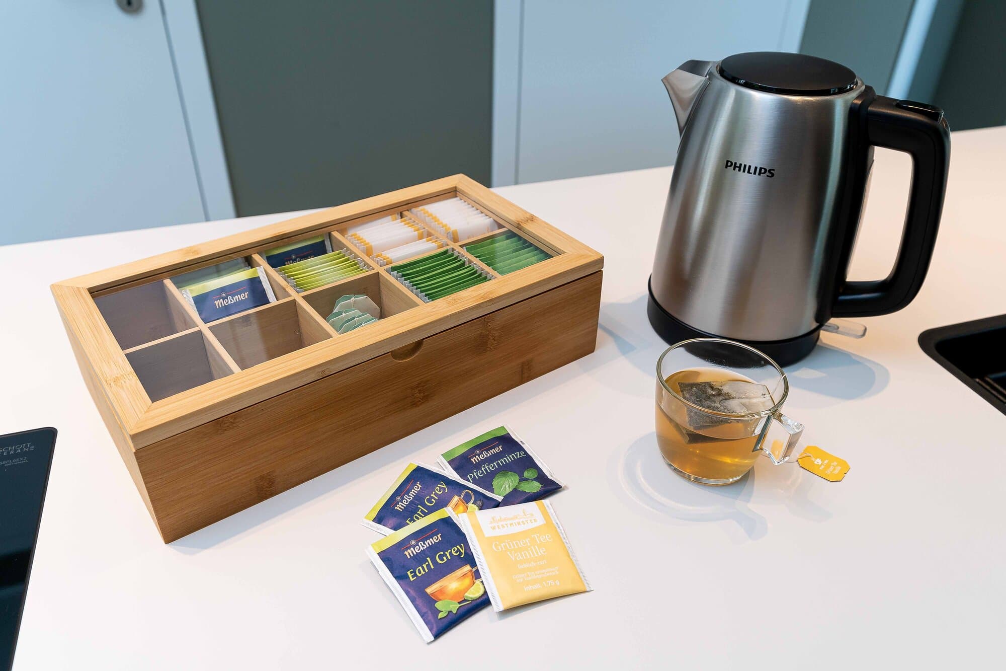 Tee-Station in der Küche, Bambusbox mit sortierten Meßmer-Teesorten, Philips Wasserkocher und eine gefüllte Glastasse